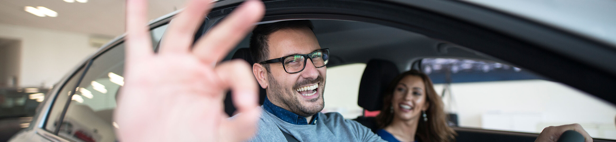 Im Vordergrund Fahrlehrer, der positiv Geste zeigt, im Hintergrund lächelnde Fahrschülerin, beide im Auto